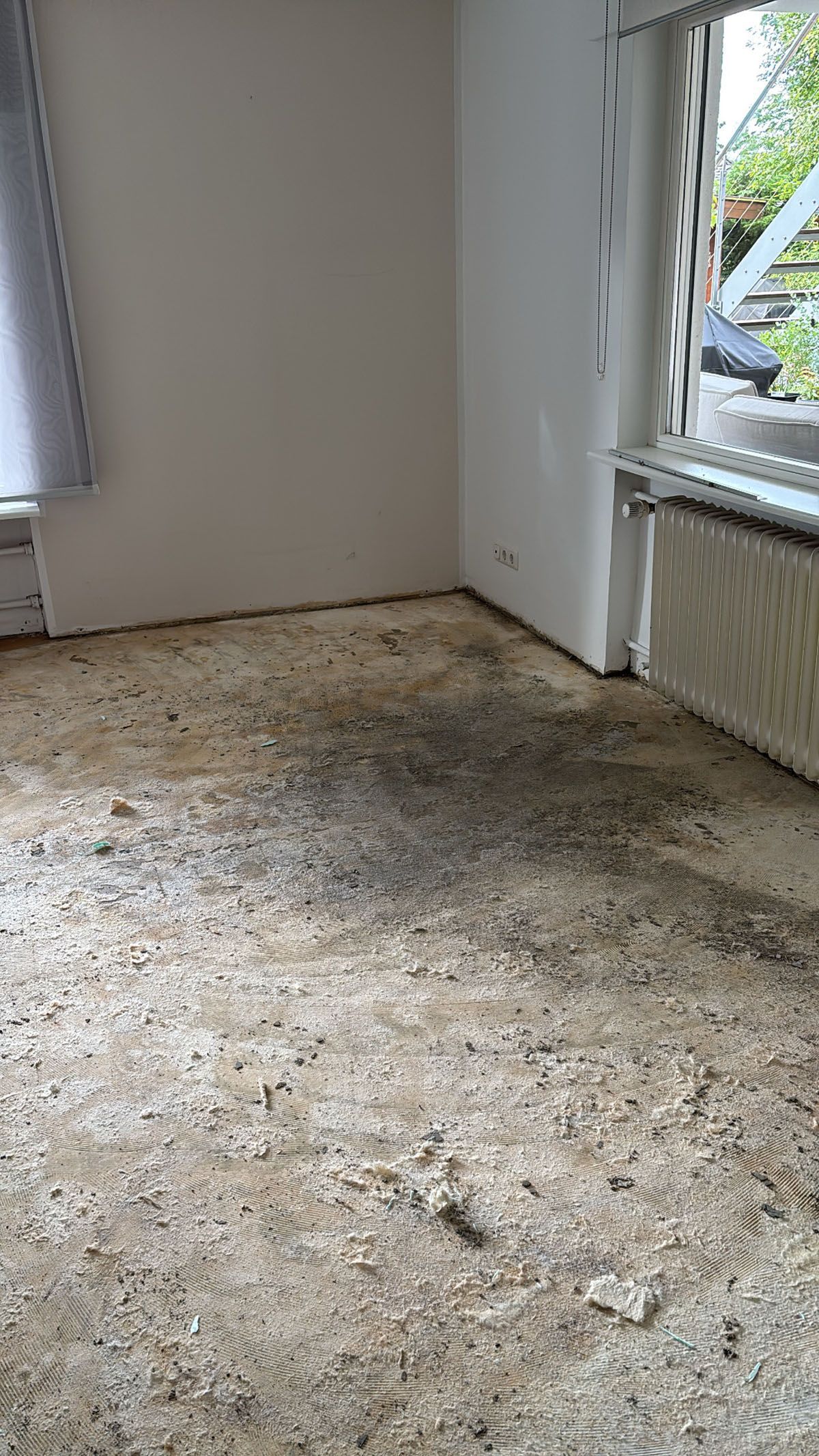 Nackter, mit Schutt bedeckter Boden in einem Raum mit Fenster, Heizkörper und weißen Wänden.