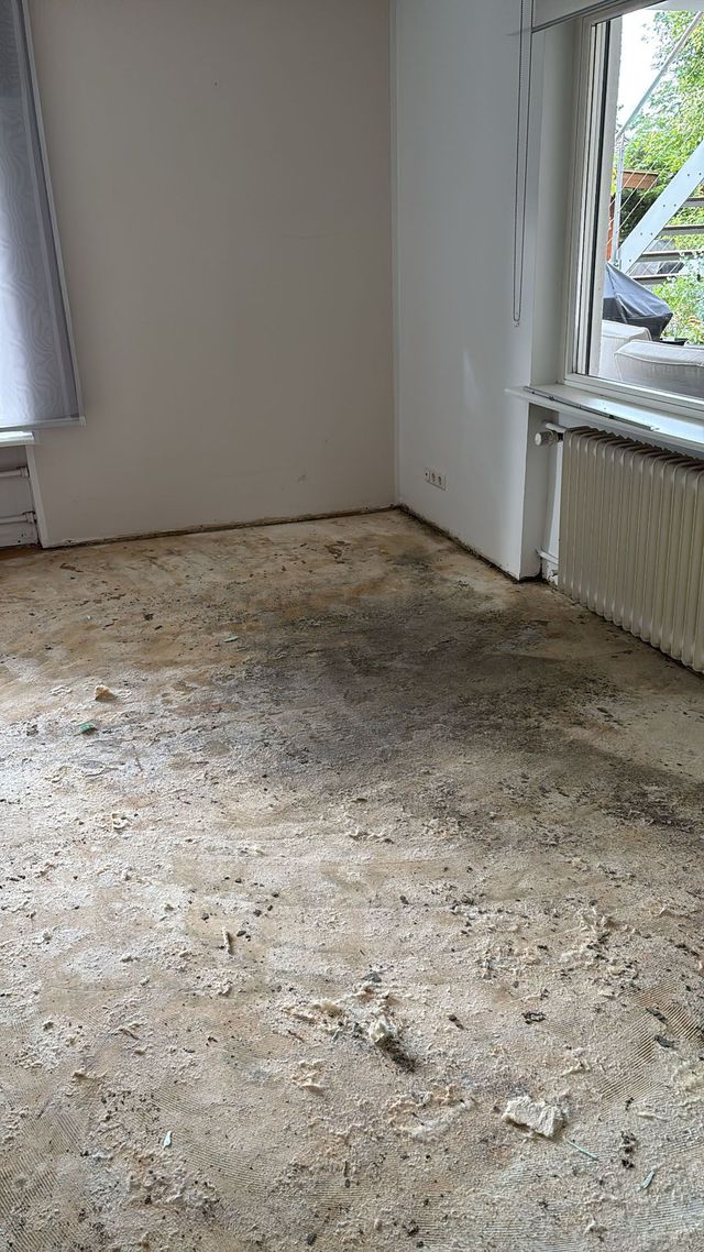 Rohboden mit Resten des Bodenbelags, neben einem Fenster und einem Heizkörper; Innenraum.