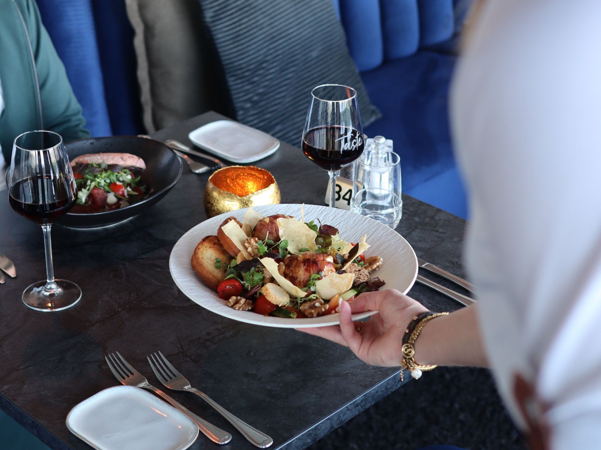 Een ober zet een salade op tafel terwijl twee mensen in een restaurant eten. Er wordt rode wijn geserveerd.