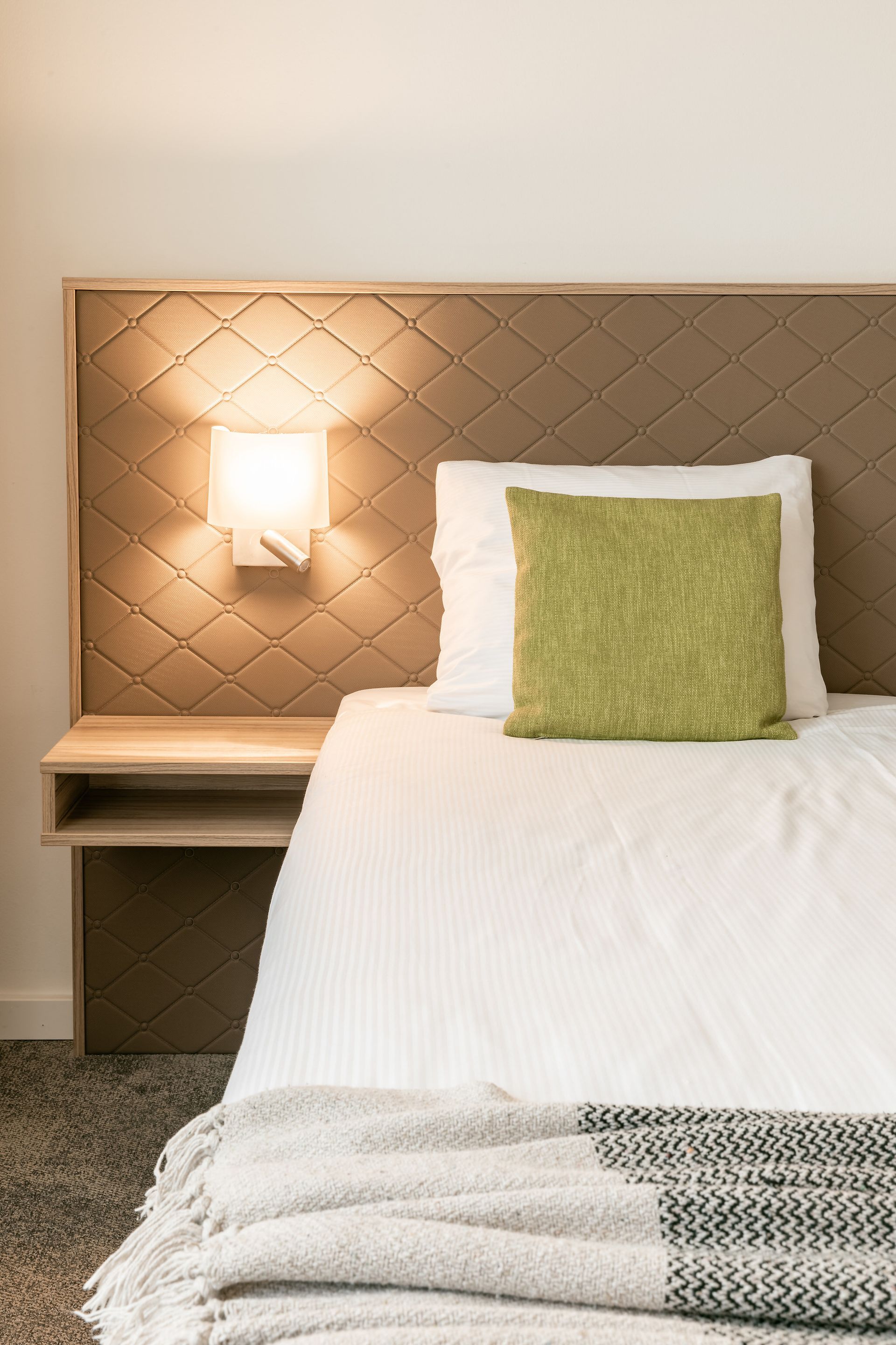 Interieur kamer Churchill Hotel Terneuzen. Bed met wit linnen, groen kussen, lichtbruin hoofdeinde en een leeslamp.