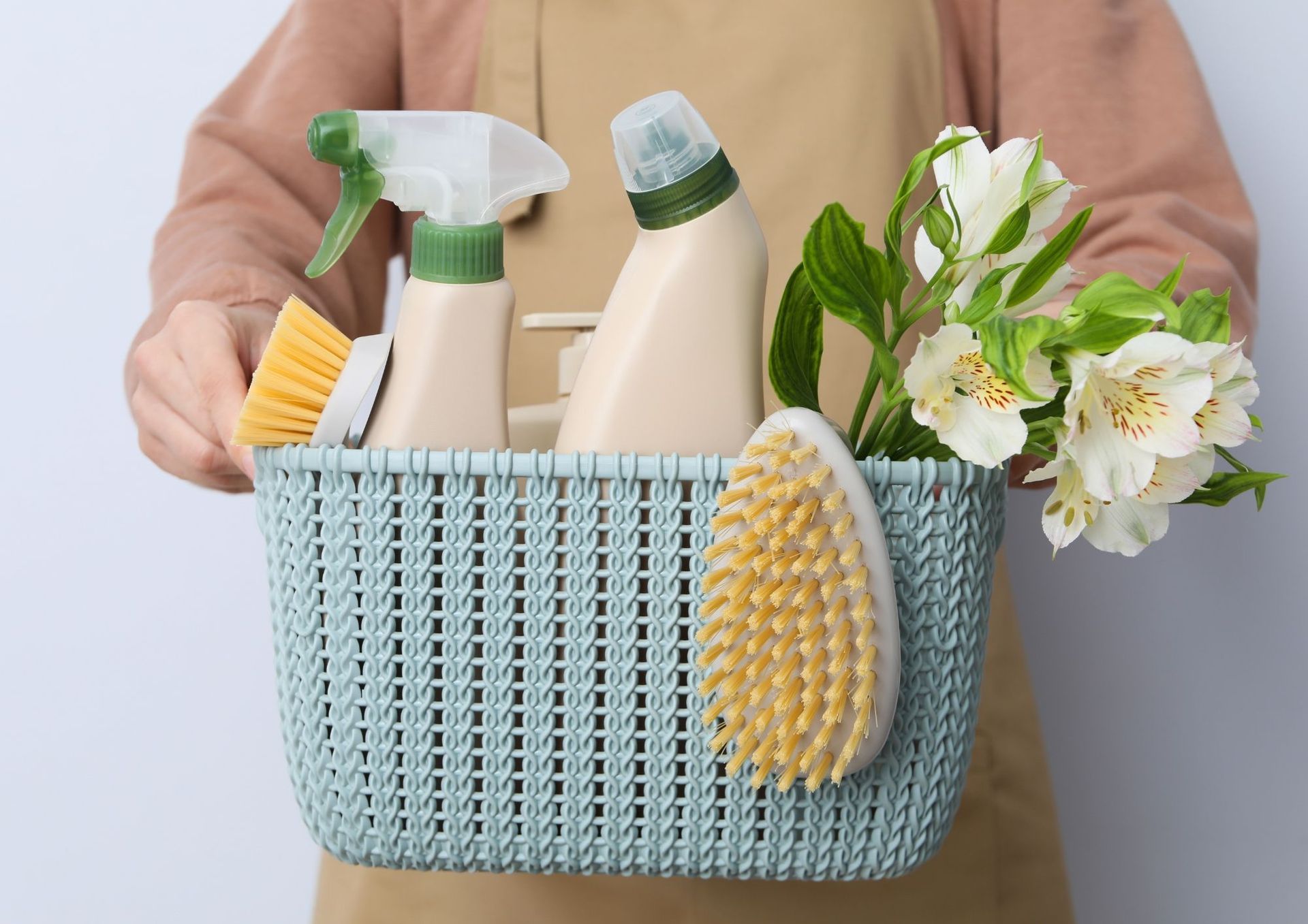Persoon die een lichtblauwe mand vasthoudt met schoonmaakmiddelen en witte bloemen.