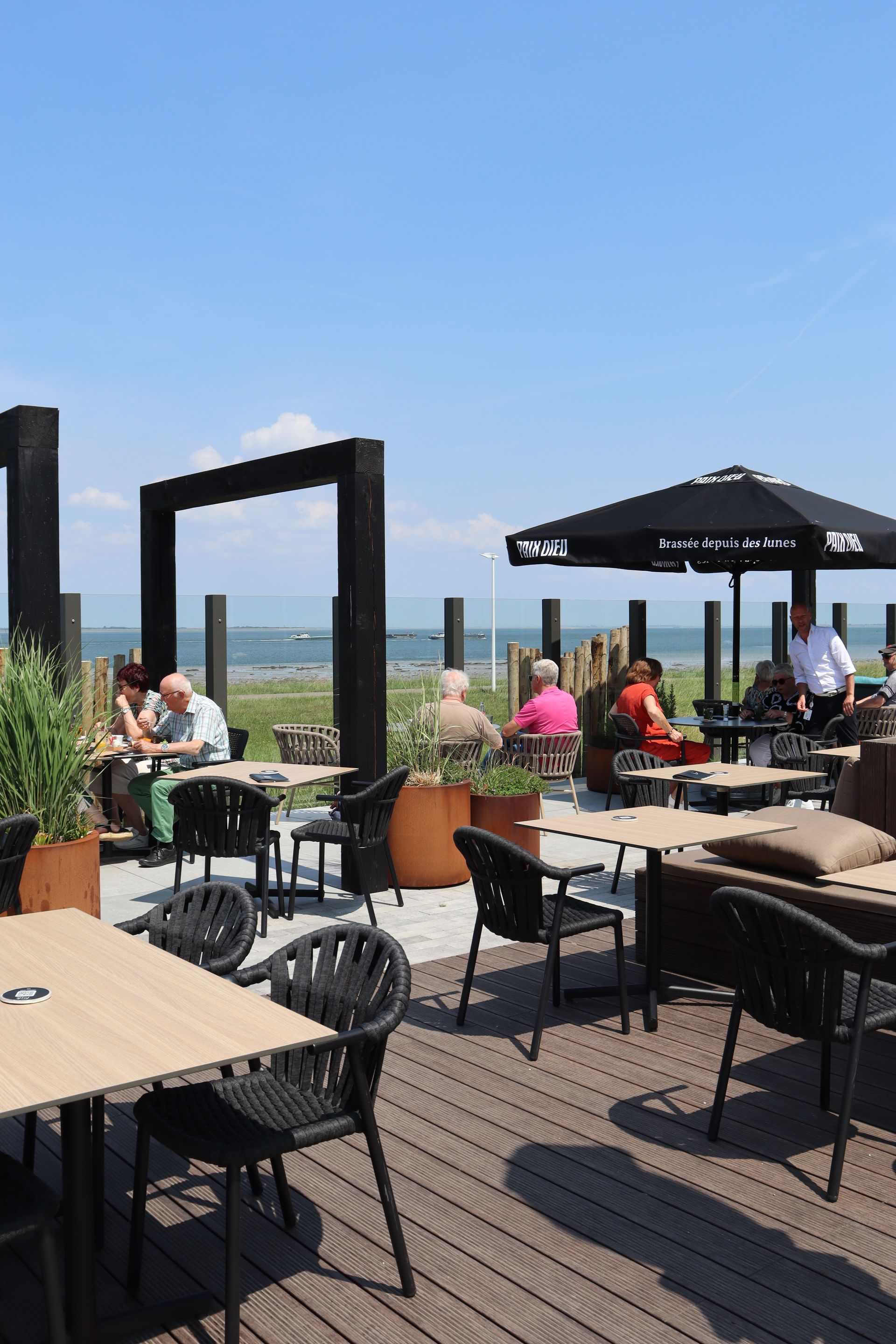 Outdoor restaurant patio with tables, chairs, and customers dining under umbrellas on a sunny day.