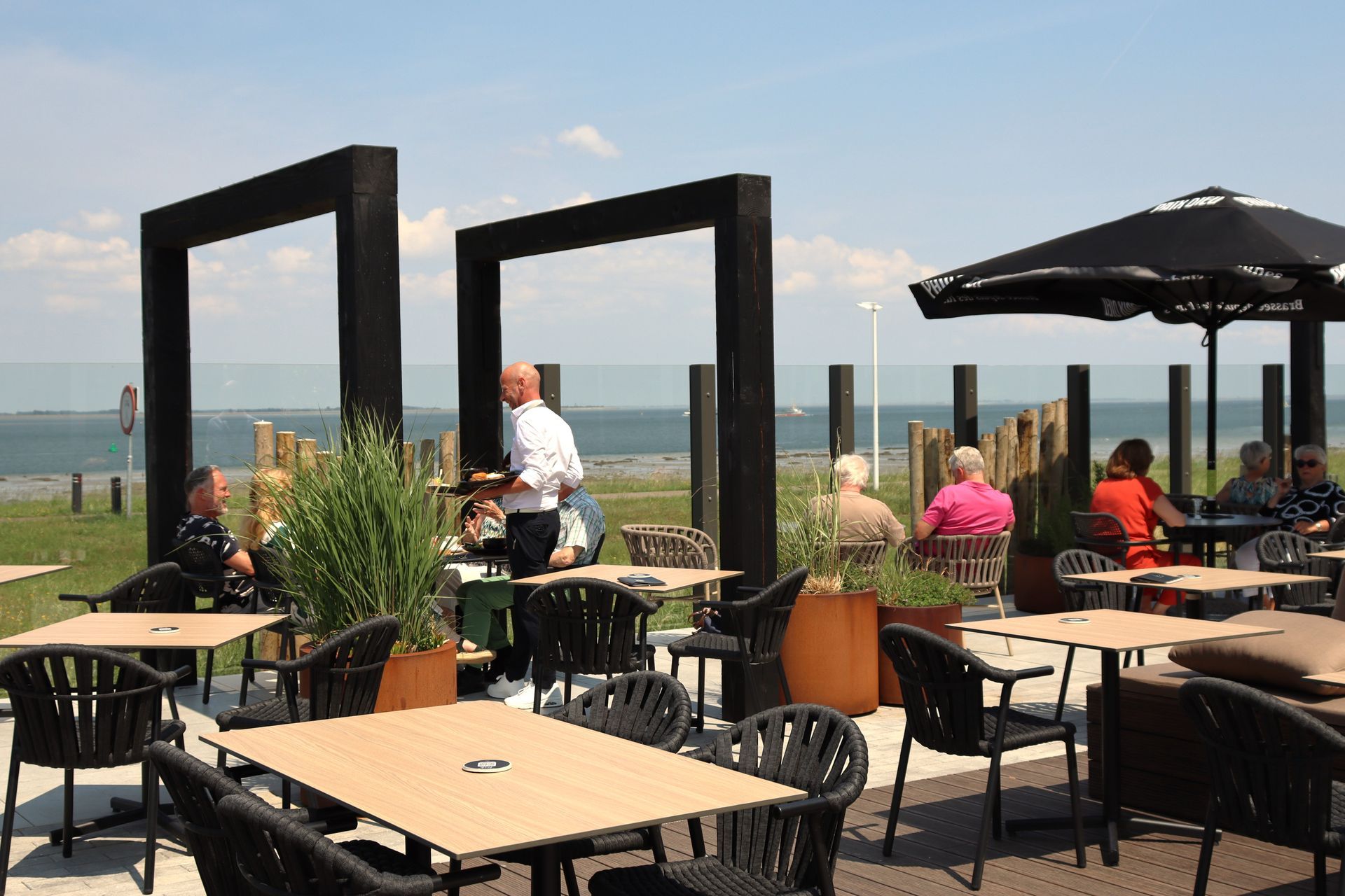 Outdoor cafe: diners at tables, waiter serving, ocean view under blue sky.
