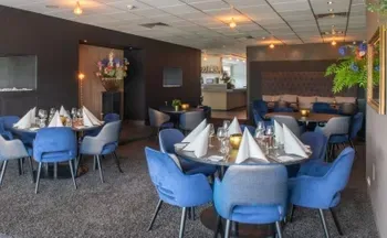 Interieur van restaurant met ronde tafels om te dineren, blauwe stoelen, donkere muren en een bar op de achtergrond.