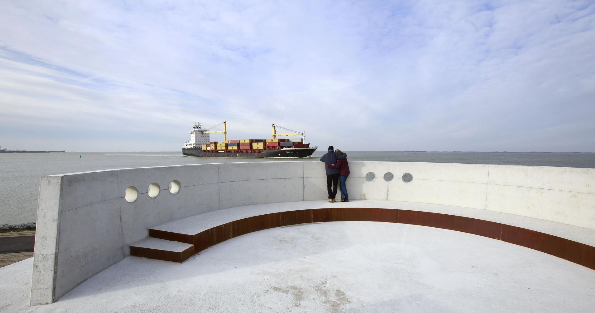Twee mensen op een betonnen observatiedek kijken naar een vrachtschip op het water. Bewolkte lucht.