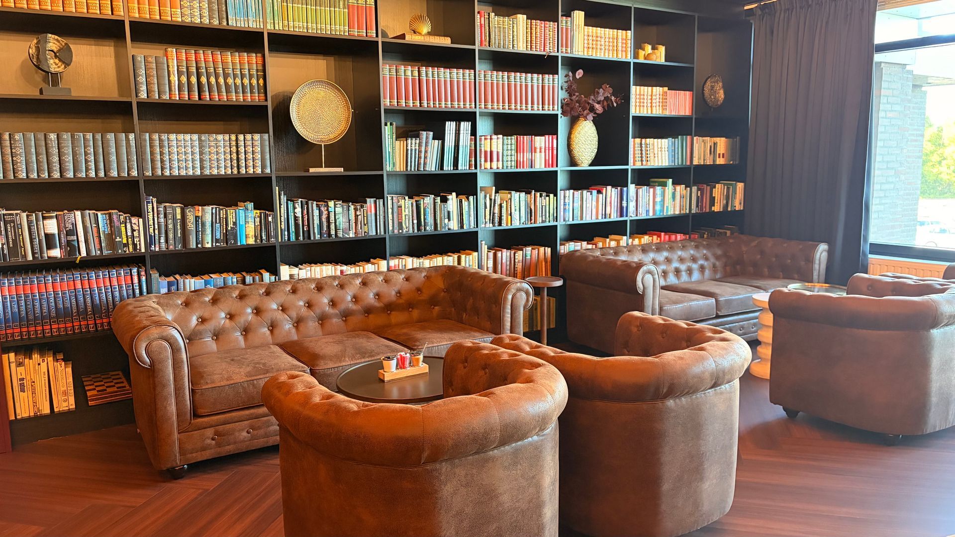 Cozy lounge with brown leather seating, tall bookshelves, and large windows letting in daylight