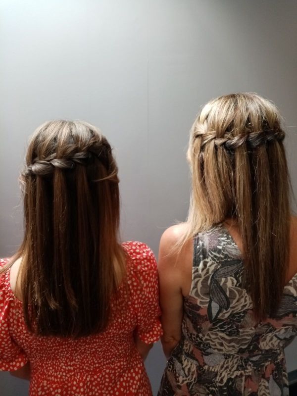 Deux femmes, de dos, aux cheveux longs et lisses. Toutes deux portent des bandeaux tressés. L'une porte une robe rouge, l'autre une robe à imprimé floral.