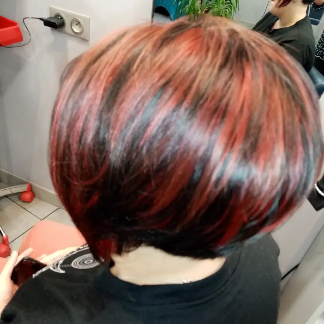 Vue arrière d'une personne avec une coiffure courte et arrondie avec des reflets rouges, noirs et brun foncé dans un salon.