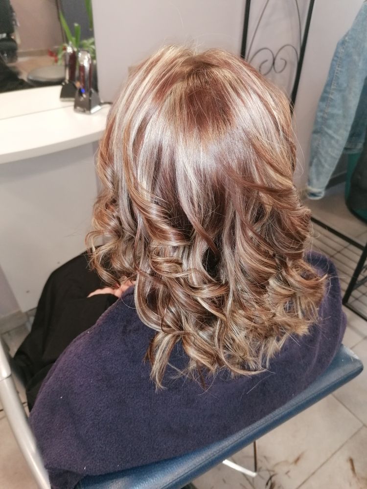 Femme aux cheveux bouclés blonds et bruns méchés, assise sur une chaise de salon.