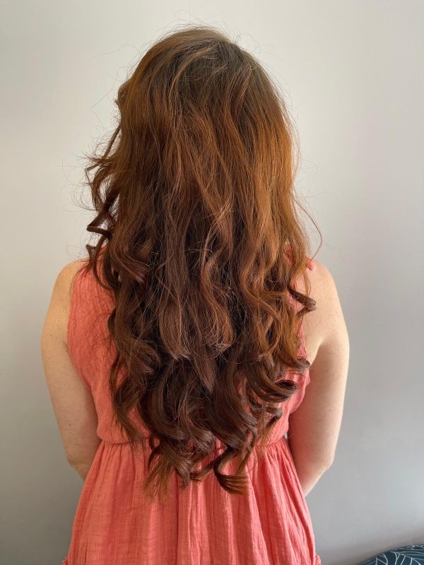 Femme aux longs cheveux auburn ondulés portant une robe couleur pêche, debout, dos à la caméra.