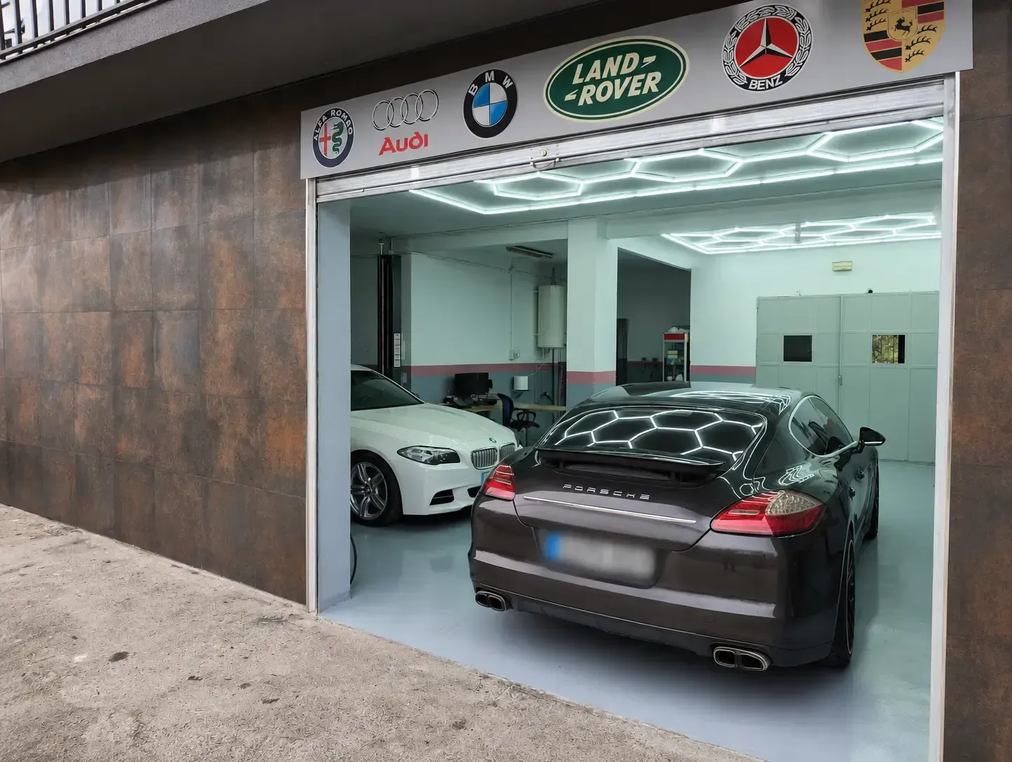 Taller de coches de lujo con dos vehículos, entre ellos un sedán negro y un BMW blanco, bajo los logotipos de las marcas.