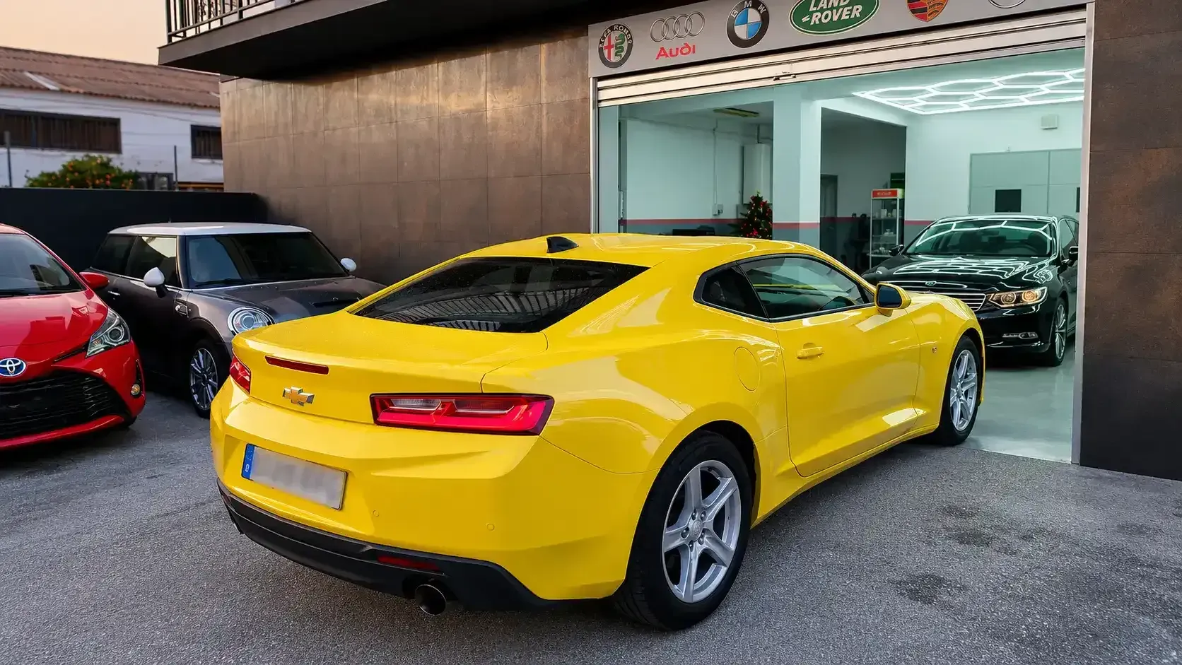Un coche deportivo amarillo aparcado frente a un concesionario de coches, con otros coches visibles en las inmediaciones.