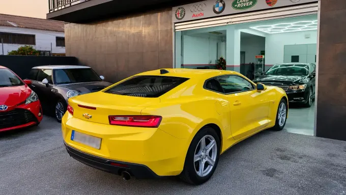 Un coche deportivo amarillo aparcado frente a un taller mecánico, con otros coches visibles en las inmediaciones.