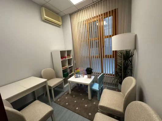 Una sala de terapia con una mesa blanca, sillas, una pequeña mesa de juegos, una estantería, una lámpara de pie