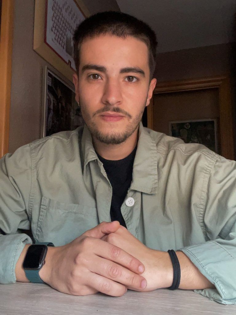 Hombre con cabello corto y oscuro, camisa verde claro sobre un top negro, manos entrelazadas, mirando hacia adelante.