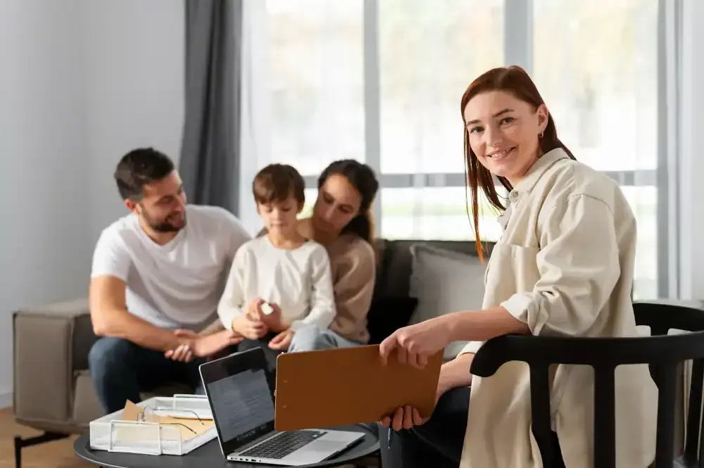 Mujer con camisa beige con su familia, ordenador portátil y portapapeles; sentada en una sala de estar. Imagen de Freepik