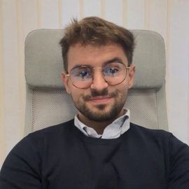 Una persona con cabello castaño corto y gafas redondas, que viste un suéter oscuro sobre una camisa con cuello