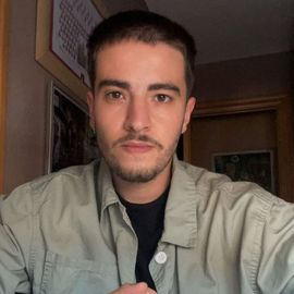 Un primer plano de una persona con cabello corto y oscuro y barba clara, que lleva una camisa beige