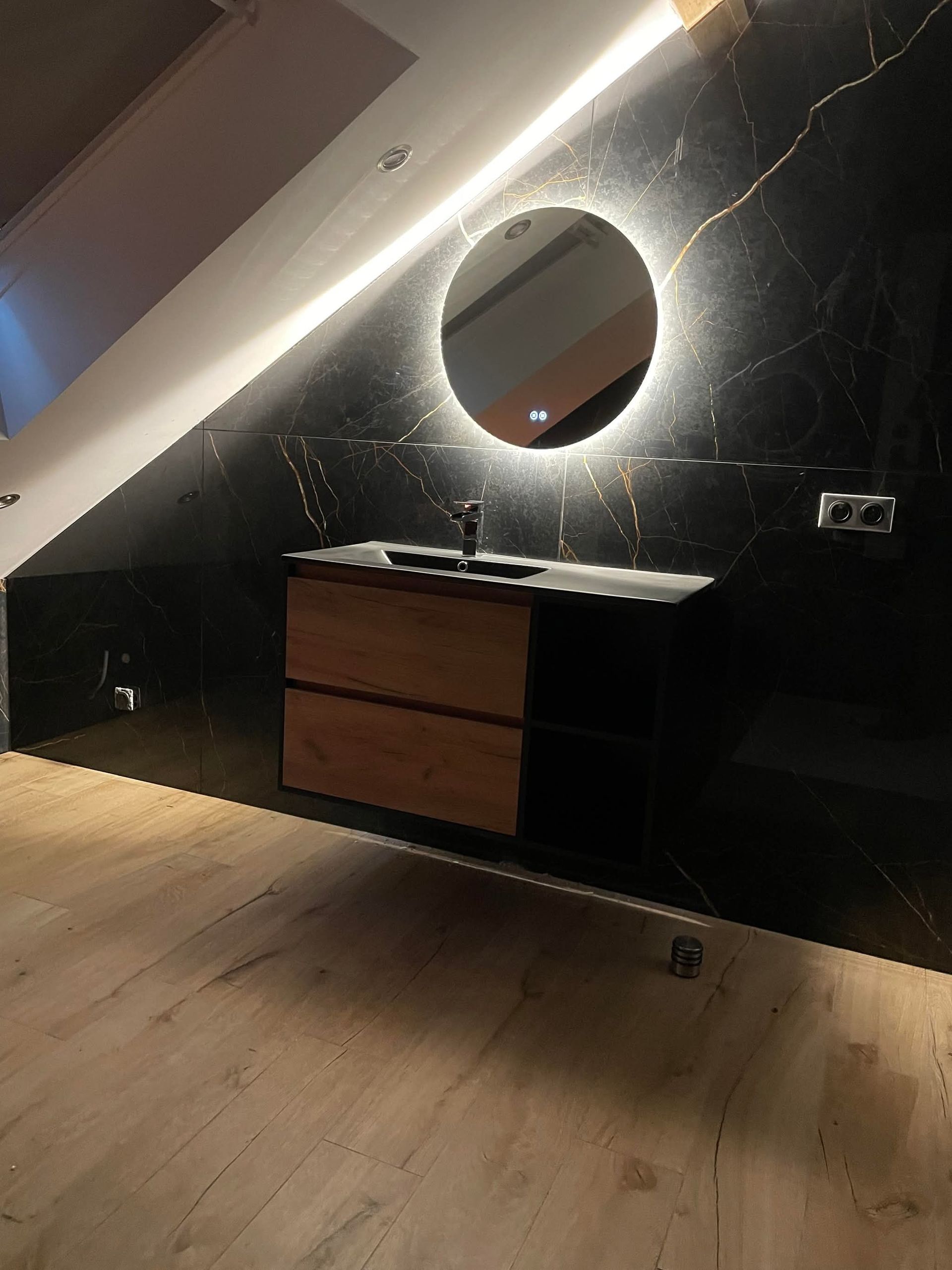 Salle de bains avec mur en marbre noir, meuble-lavabo en bois, miroir rond rétroéclairé et plafond mansardé.