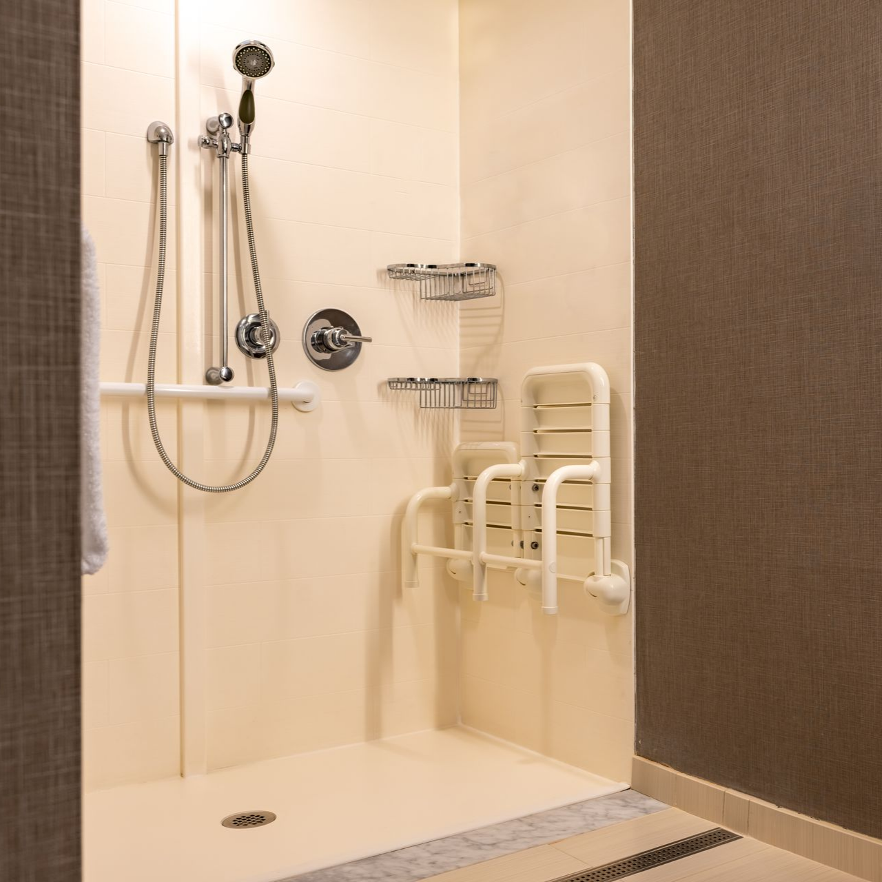 Douche accessible avec siège rabattable, barres d'appui et plusieurs étagères. Murs couleur crème et mur d'accent brun foncé.