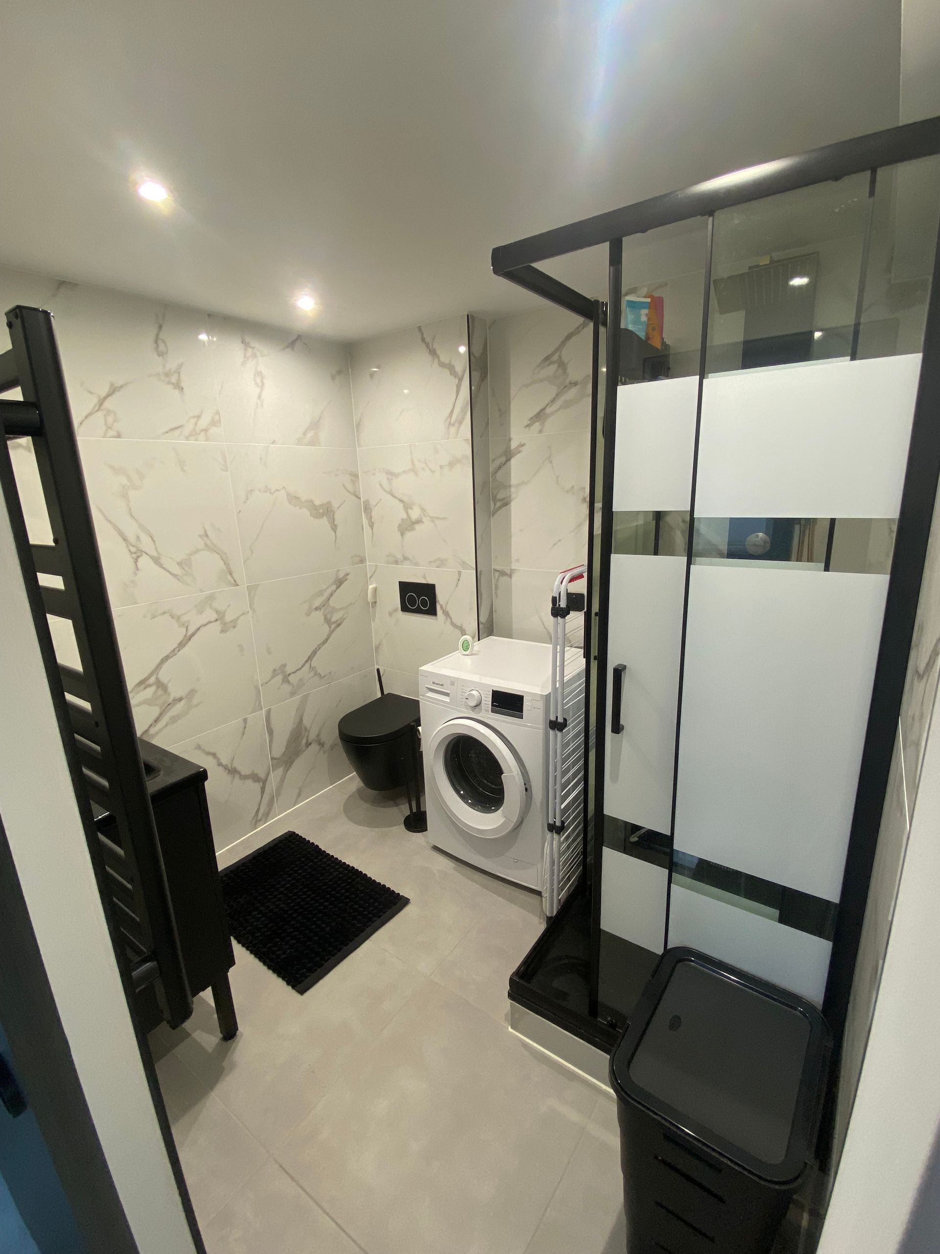 Salle de bains moderne avec carrelage blanc imitation marbre, robinetterie noire, douche, toilettes et lave-linge.