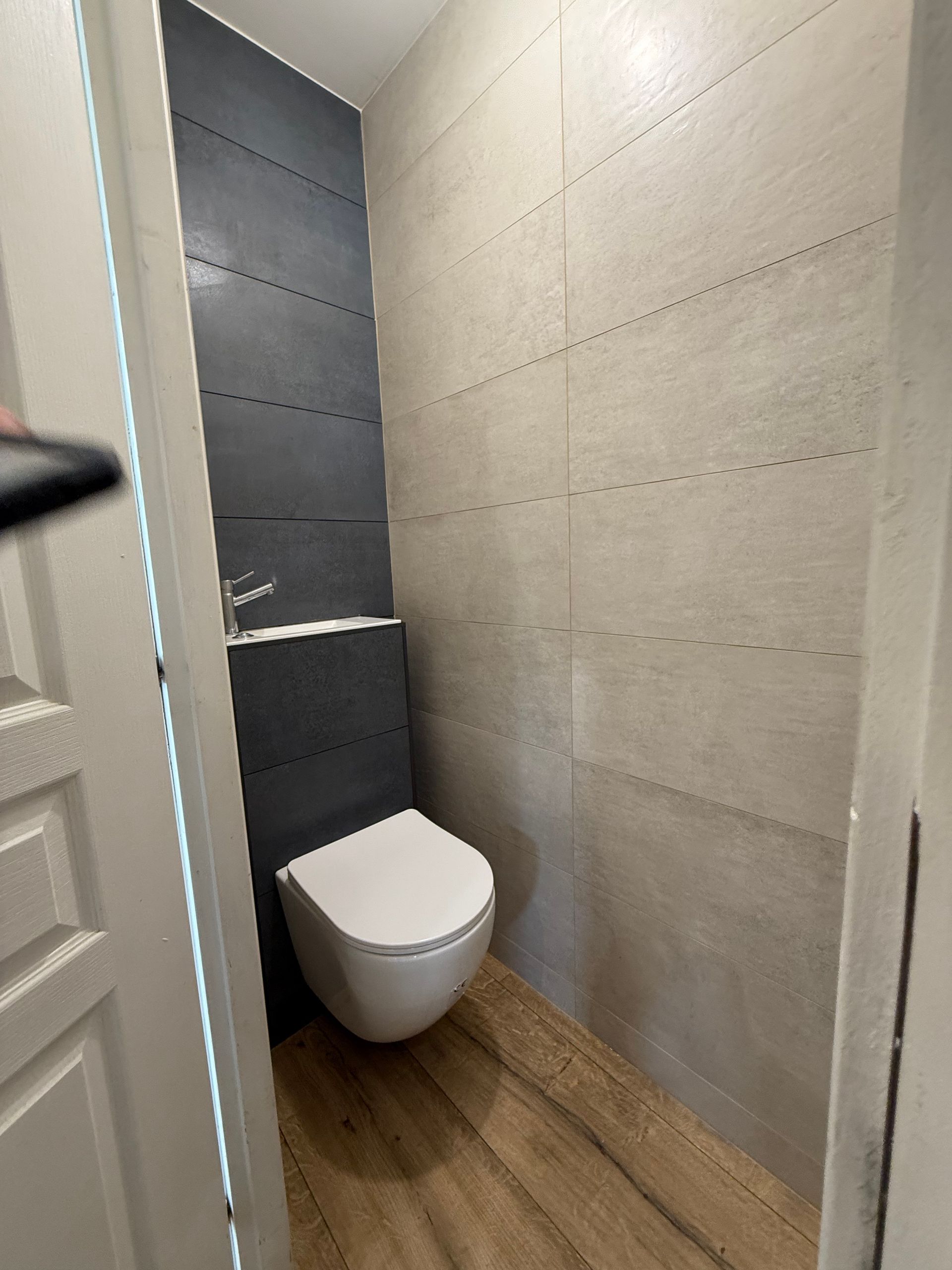 Toilettes dans une petite salle de bains aux murs carrelés gris et beiges et au sol en bois.