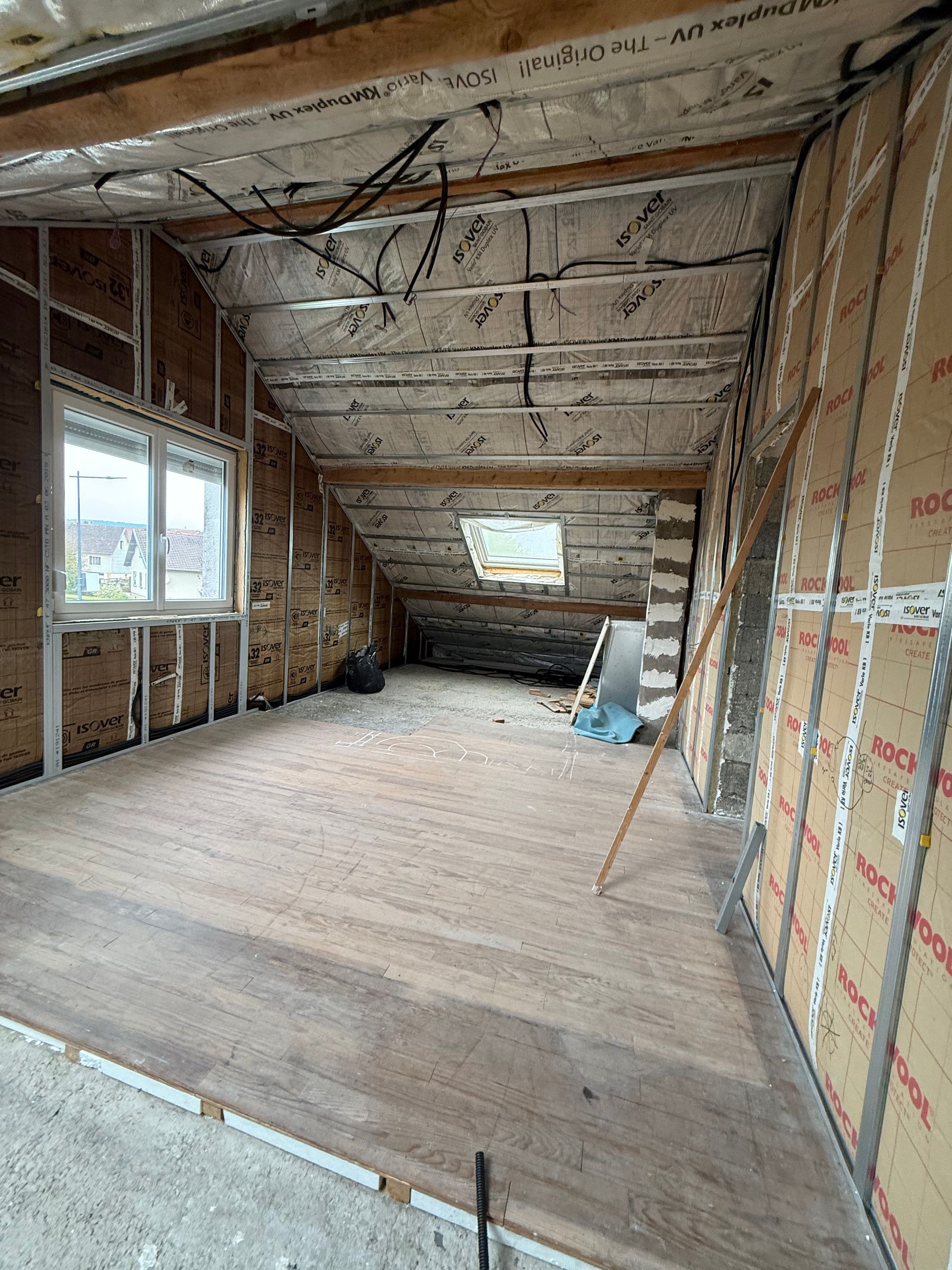 Intérieur d'un grenier en construction ; poutres apparentes, isolation, fenêtre et plancher non fini.