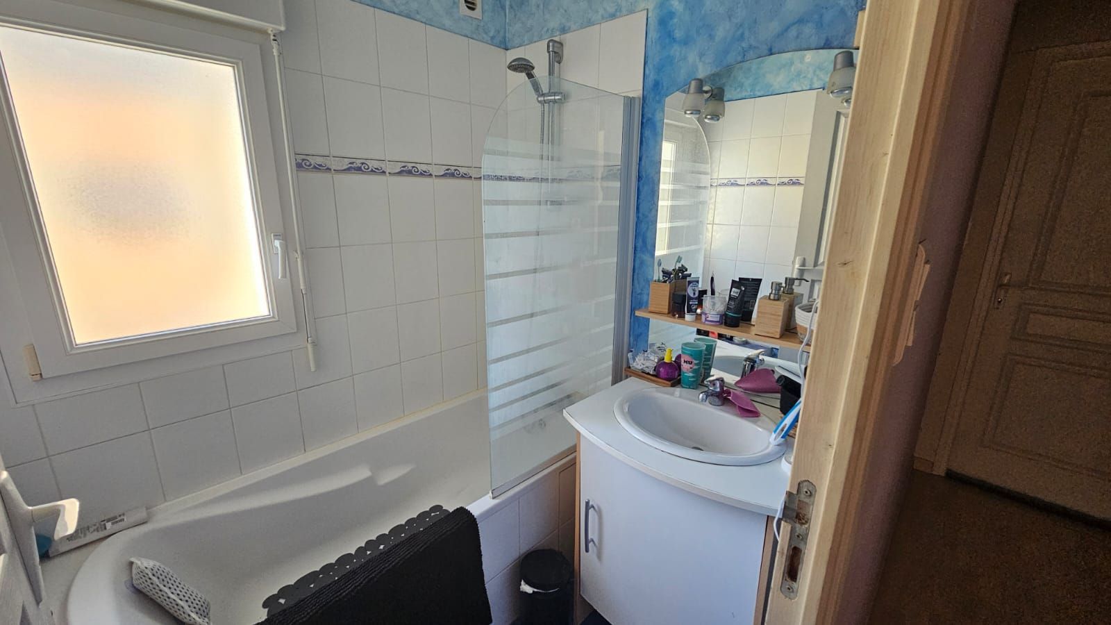 Salle de bains avec baignoire, douche, lavabo, fenêtre et armoire ; tons blancs, bleus et bruns.