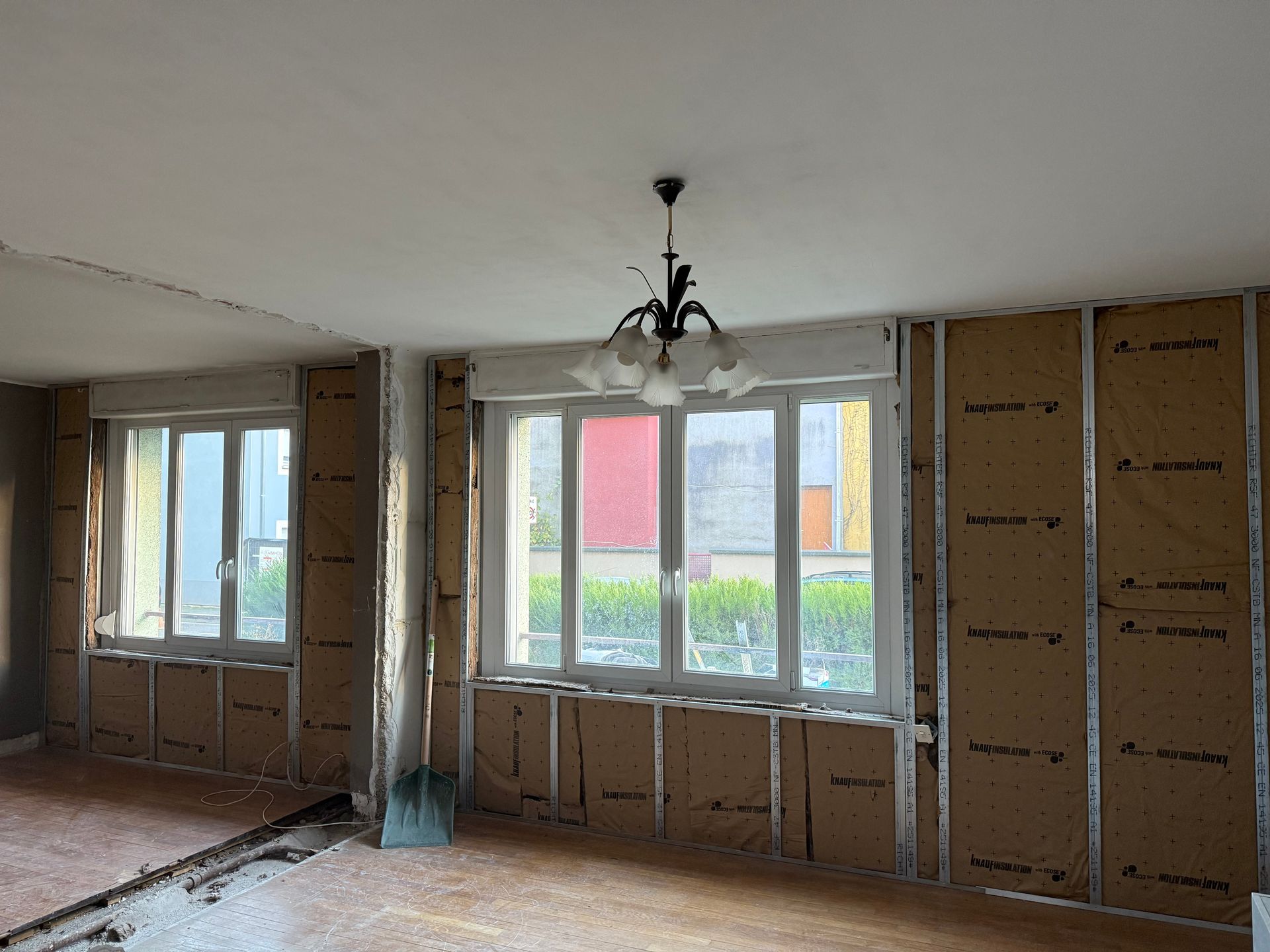 Pièce en rénovation avec montants muraux et isolation apparents ; fenêtres, lustre et pelle présents.
