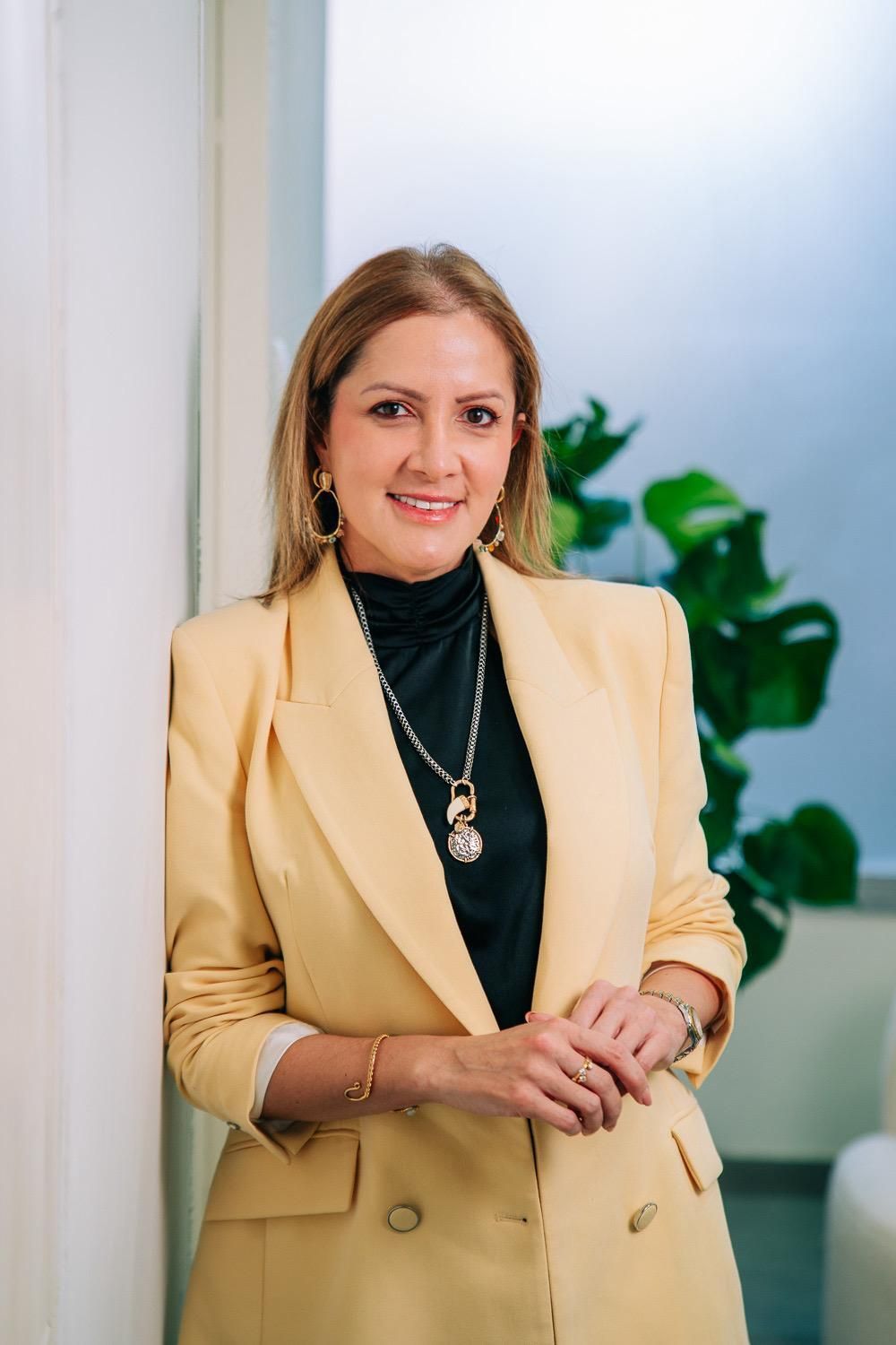 Una mujer vestida con un blazer amarillo sonríe, apoyada contra una pared, mirando a la cámara.