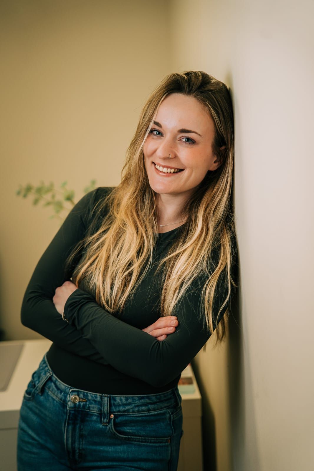 Mujer con cabello largo sonríe, se apoya contra una pared, con los brazos cruzados.