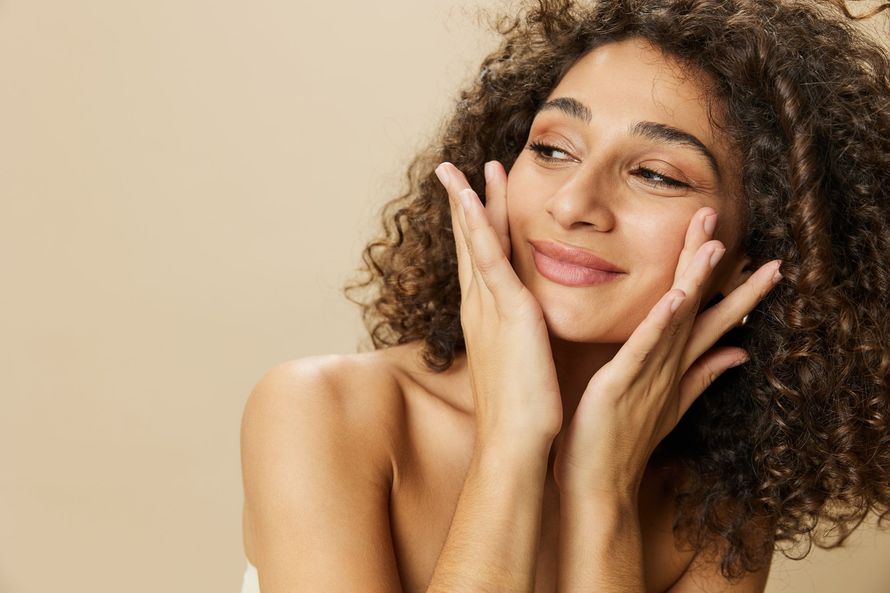Mujer con cabello rizado, manos en las mejillas, sonriendo sobre un fondo beige claro.