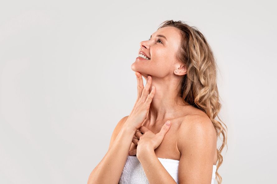 Mujer con cabello rubio y rizado se toca el cuello y el pecho, sonriendo hacia arriba, envuelta en una toalla.