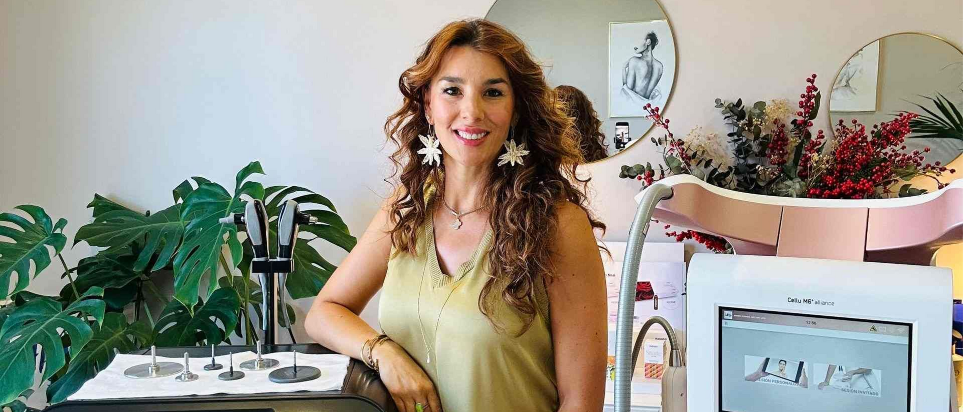 Mujer con cabello rizado sonríe, parada junto a un dispositivo con pantalla, cerca de plantas.