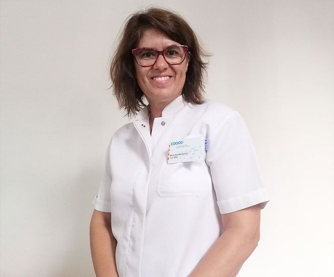 Mujer con bata blanca, gafas, sonriendo; en el interior, etiqueta con nombre visible.