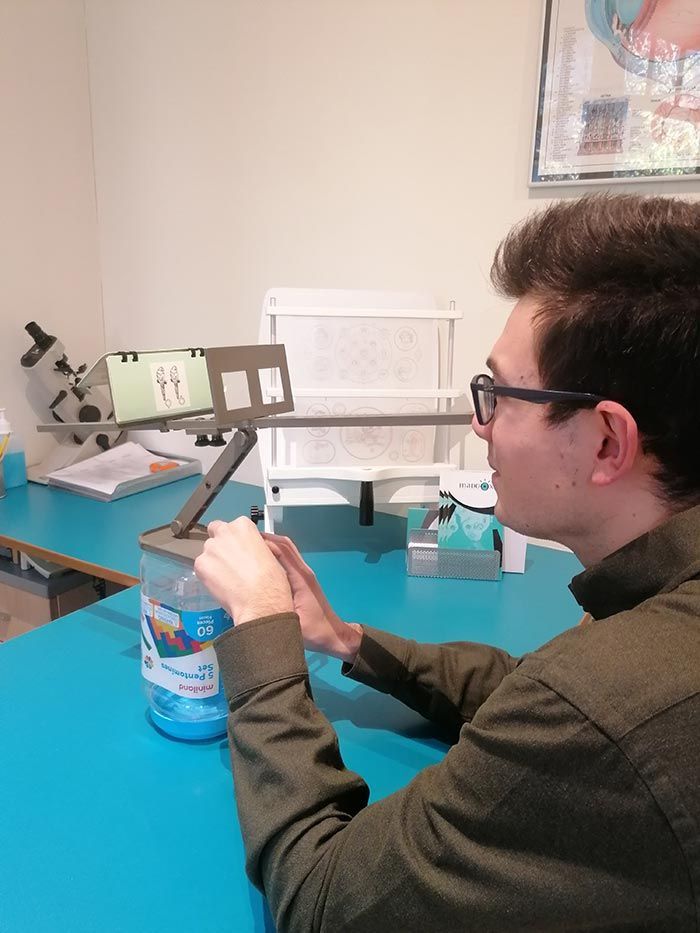 Un hombre sostiene un frasco y mira una tableta en un soporte. Está sentado en una mesa azul en una habitación.