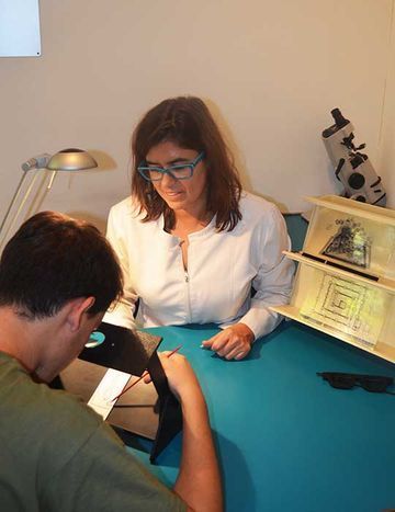 Una mujer con gafas se inclina sobre una mesa con una persona; una pequeña luz ilumina la escena, que contiene una caja de madera con arte y gafas de sol.