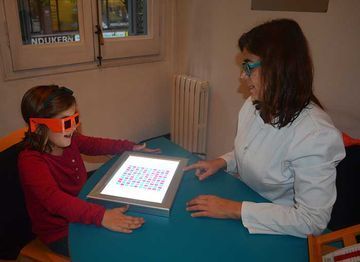 Una chica con gafas naranjas mira una pantalla iluminada mientras una mujer la señala. Están en un espacio interior.