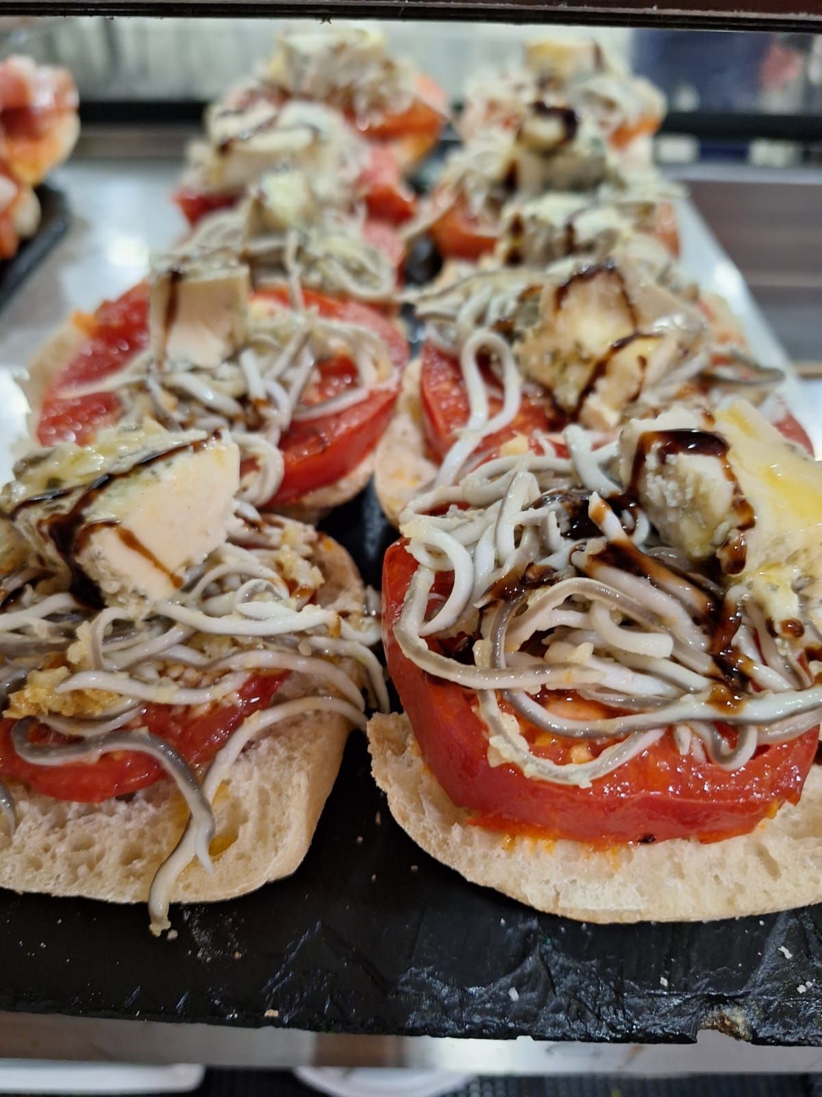 Sándwiches abiertos con tomates, anguila, queso y salsa sobre una superficie oscura.
