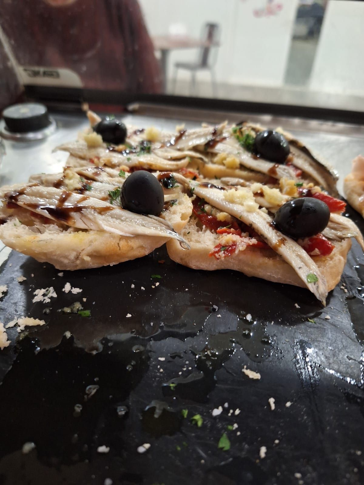 Dos sándwiches abiertos cubiertos con anchoas, aceitunas y salsa sobre una superficie negra.