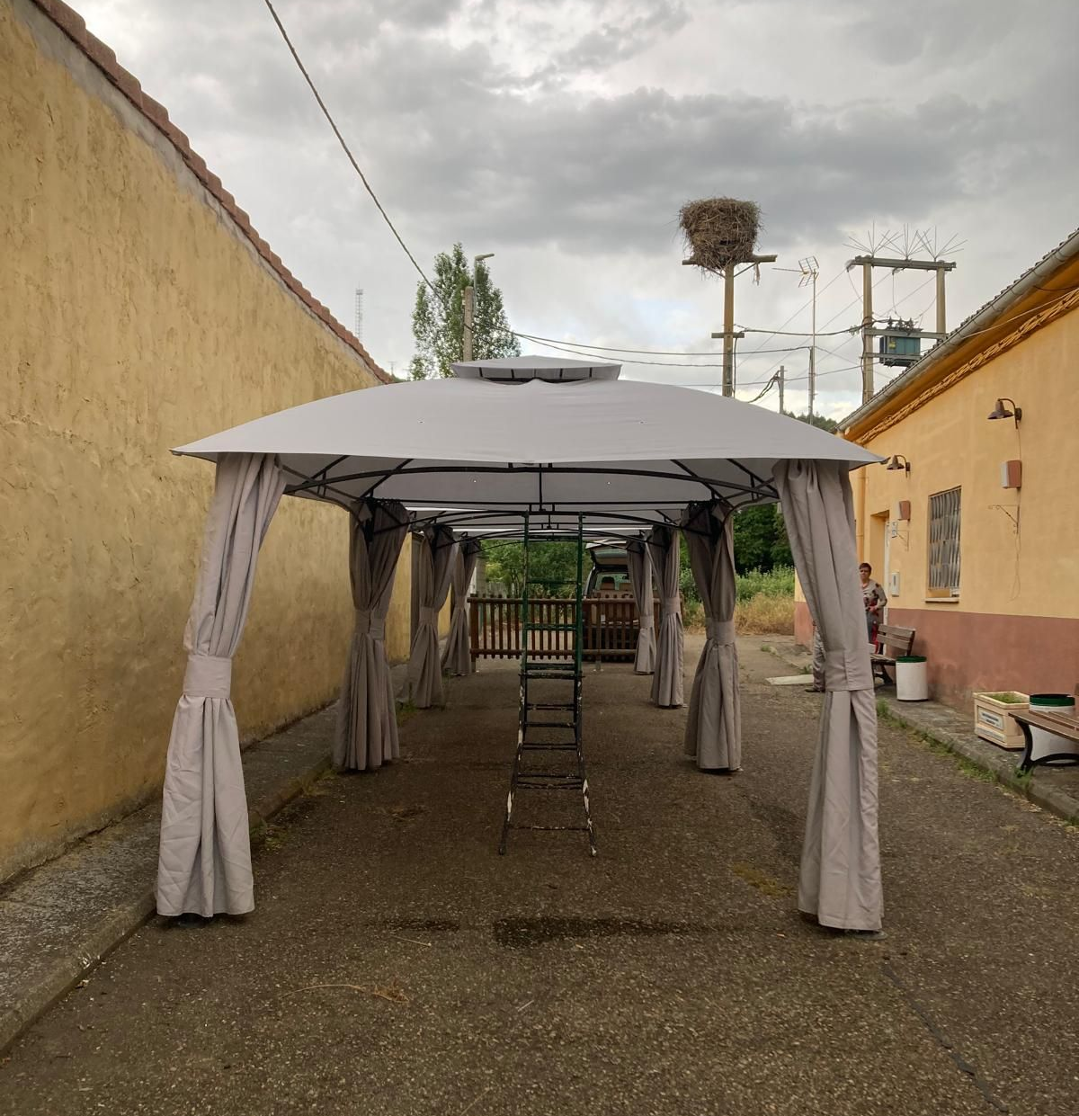 Cenador gris con cortinas, en un callejón entre edificios. Anidado sobre un poste de electricidad al fondo.