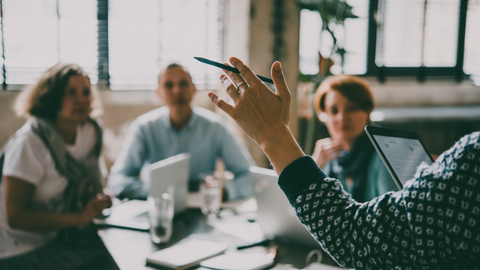 A person gestures with a pen during a collaborative office meeting with three colleagues sitting at a table.