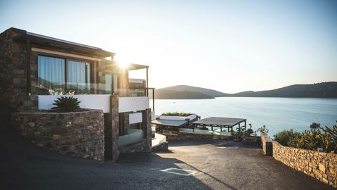 A modern stone villa overlooking a calm sea at sunset, with a paved road leading towards the water.