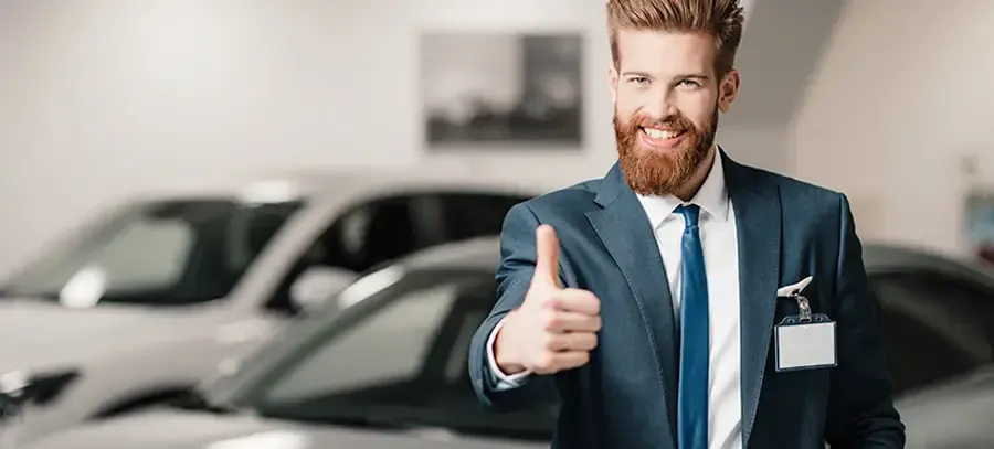 Un hombre con traje y corbata hace un gesto de aprobación con el pulgar delante de un coche.