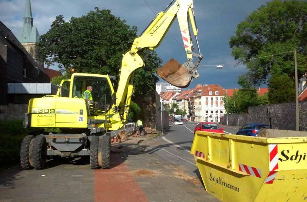Ein gelber Bagger lädt Erde in einen gelben Müllcontainer.