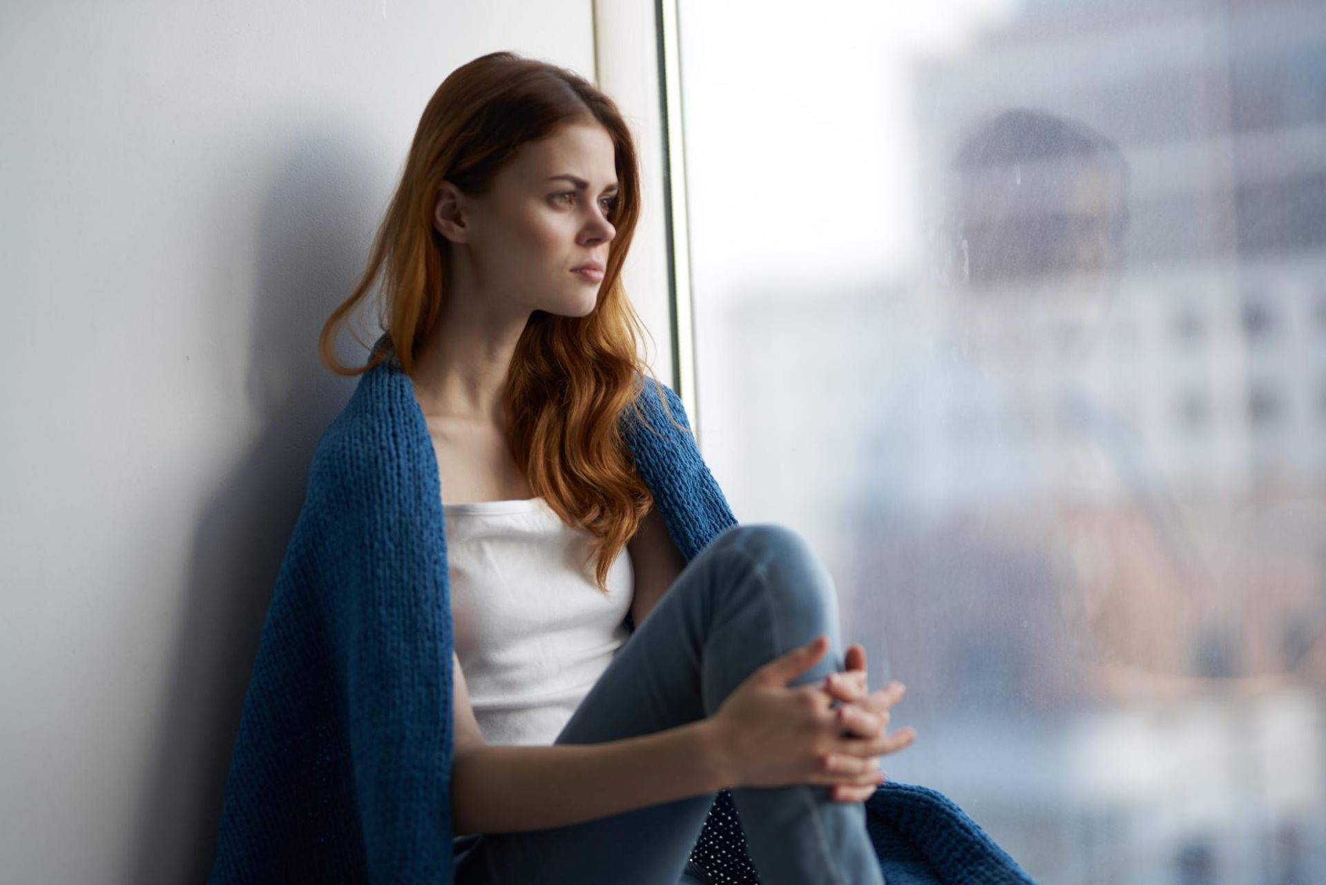 Mujer sentada junto a una ventana, con un cárdigan azul, mirando pensativamente hacia afuera.