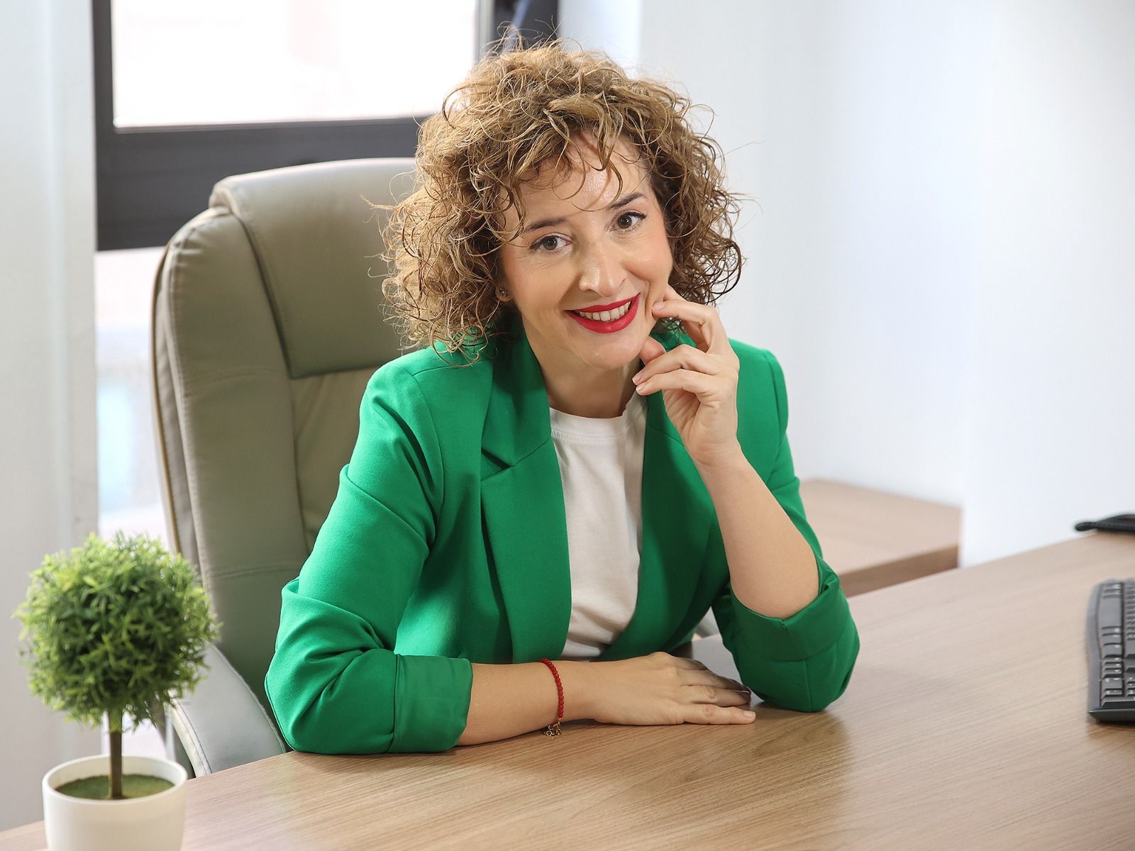 Mujer sonriente sentada en un escritorio con un blazer beige, en una oficina con una pequeña planta y un teléfono cerca.