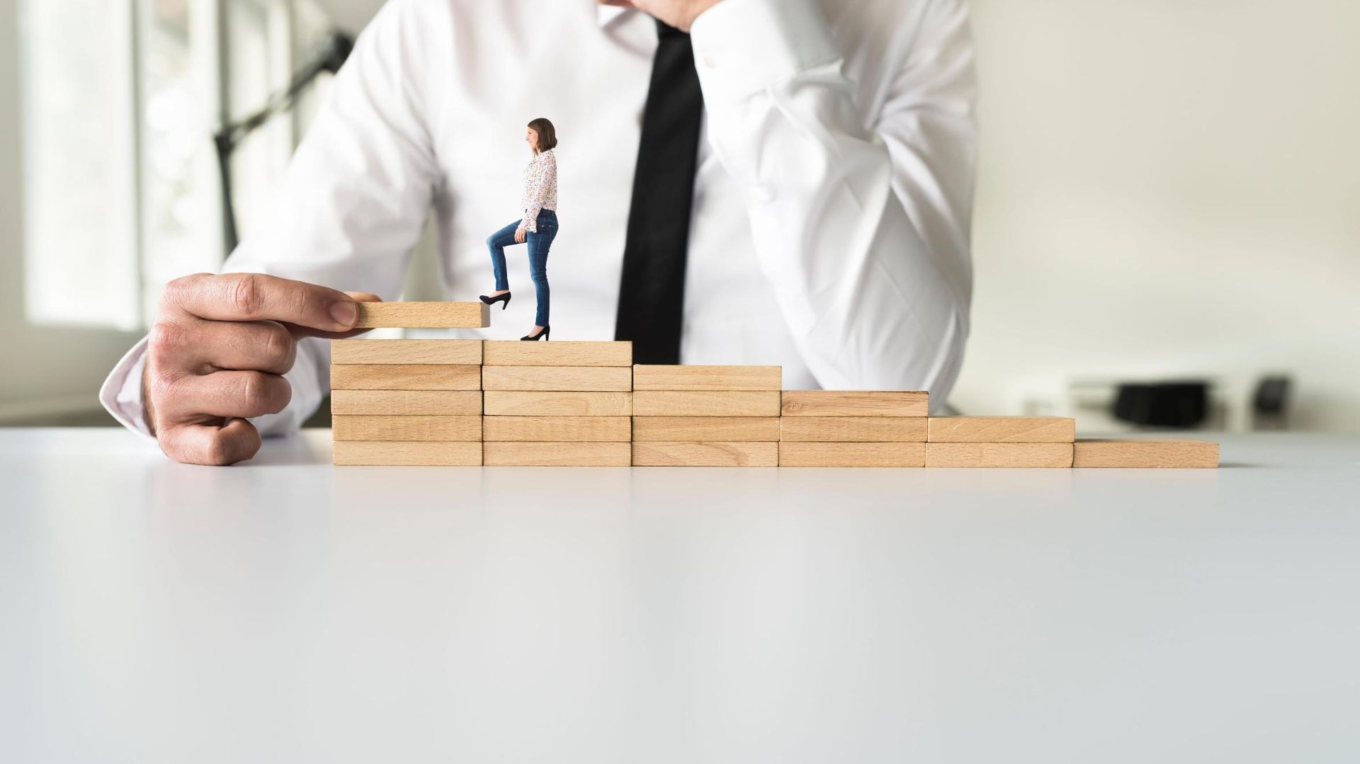 Un empresario construye un escalón con bloques de madera apilados junto a una pequeña figura sobre un escritorio.