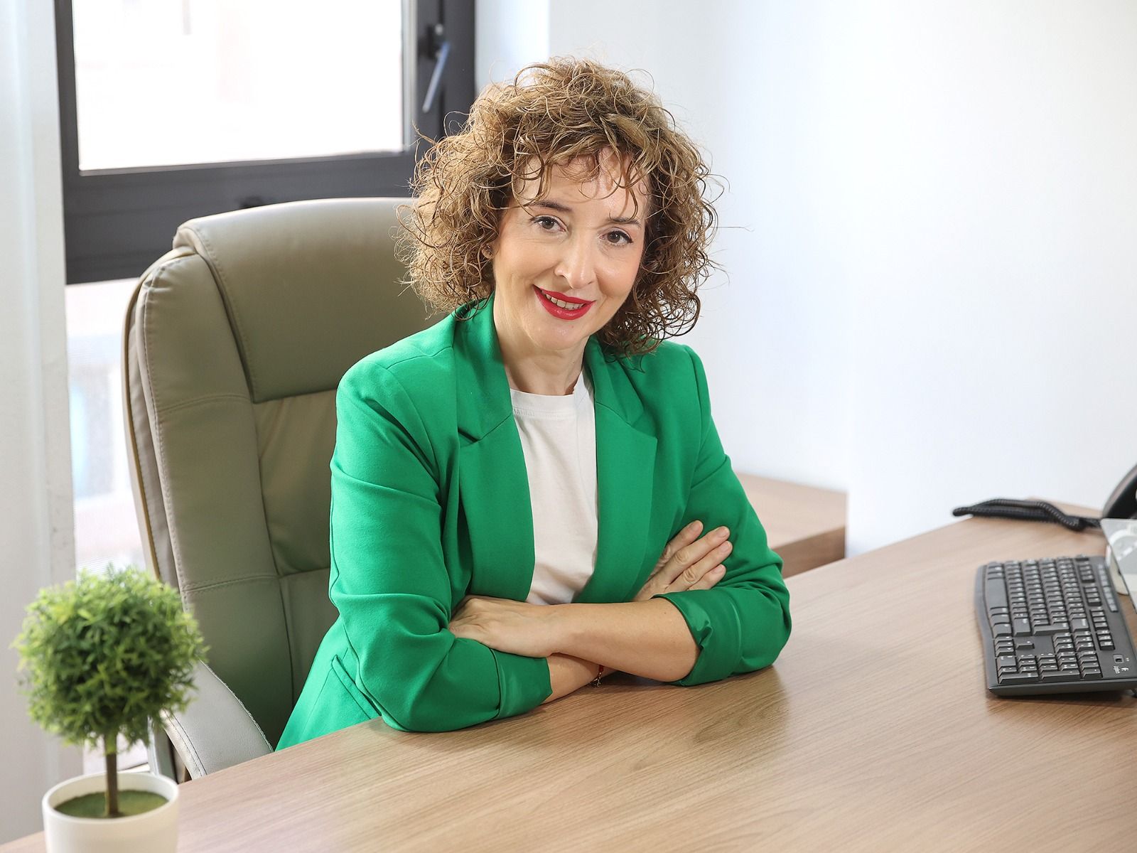 Una persona sonríe sentada en un escritorio de una oficina, con un teclado de ordenador y una pequeña planta cerca.