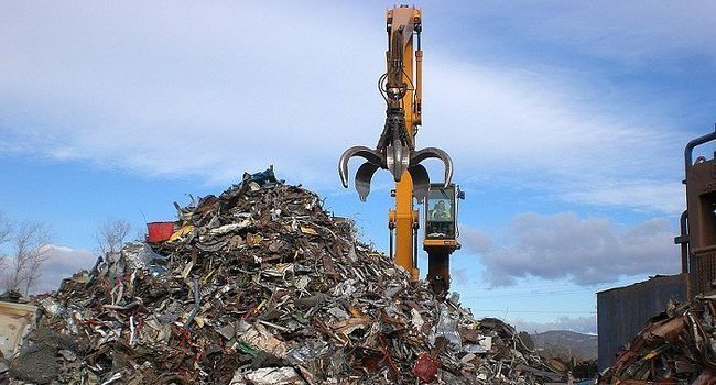Una excavadora con garra recoge chatarra de una gran pila en un depósito de chatarra, fondo de cielo azul.